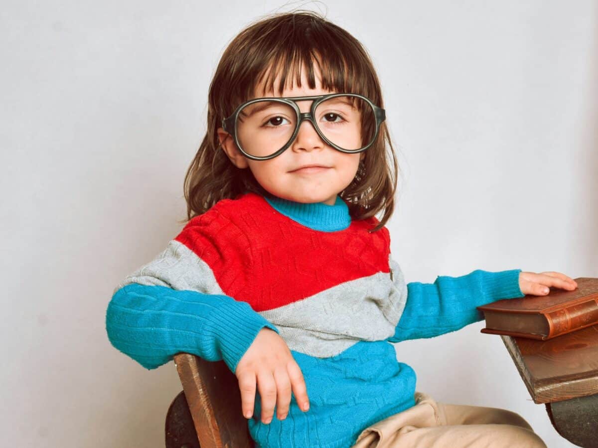 Petite fille au prénom rétro portant de grandes lunettes, assis à un vieux pupitre, avec un pull coloré et des livres posés devant elle.